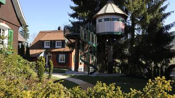 A traditional house with a red roof and green shutters, a small tower with a spiral staircase, and a well-maintained garden.