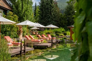 Un área de piscina tranquila al aire libre con sillones reclinables y sombrillas, rodeada de exuberante vegetación y montañas al fondo.