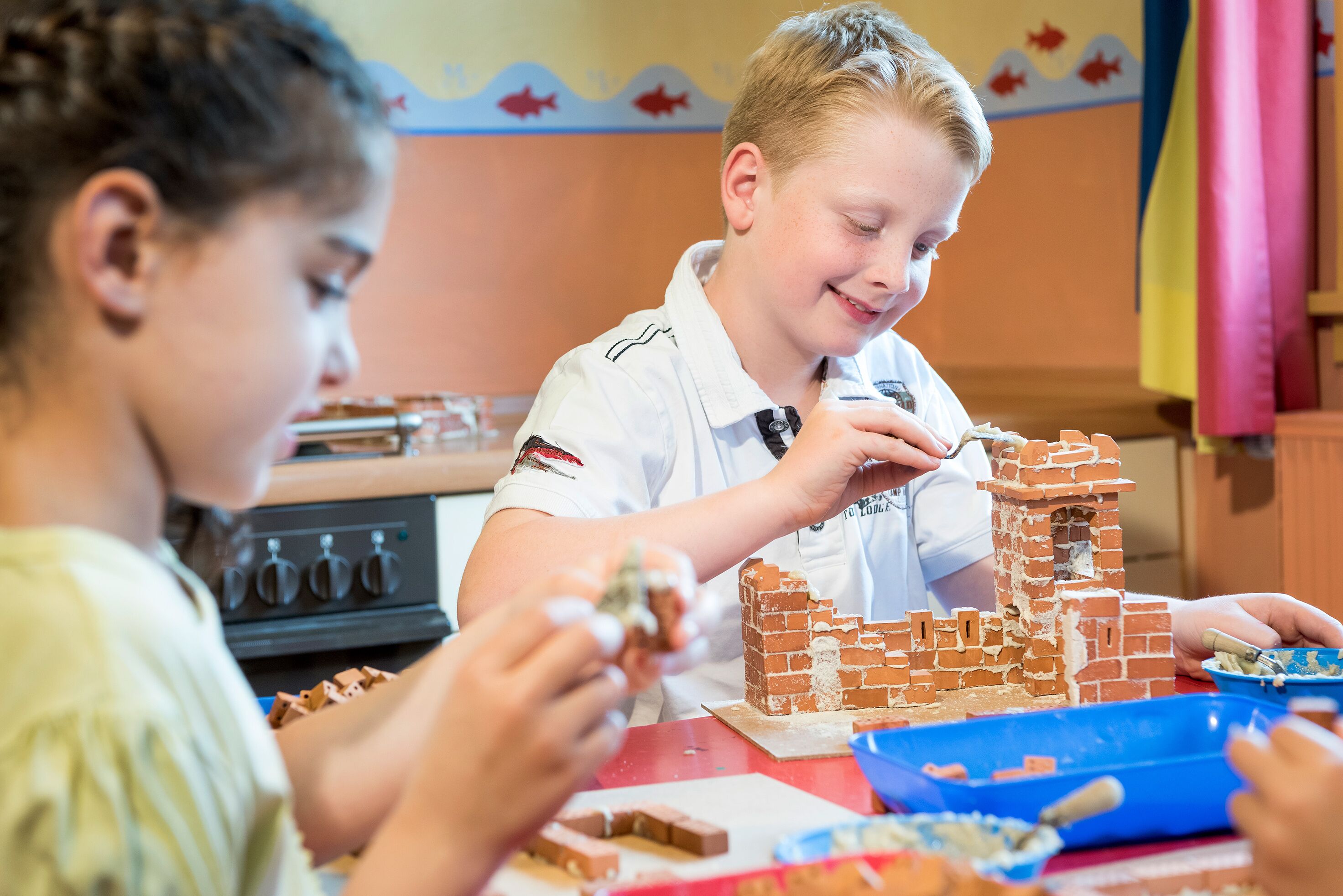 Children engaged in building a brick structure, possibly a model castle, with various building materials on a table.