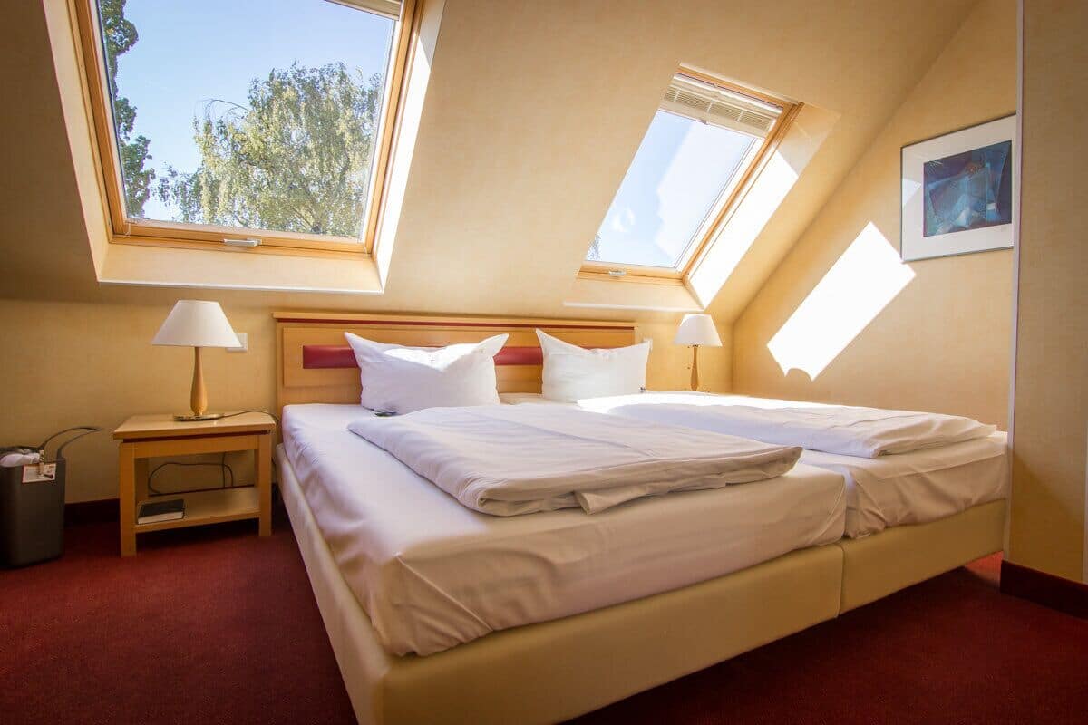 Quarto com uma cama grande, duas mesas de cabeceira, uma luminária, um cesto de lixo e janelas no teto.