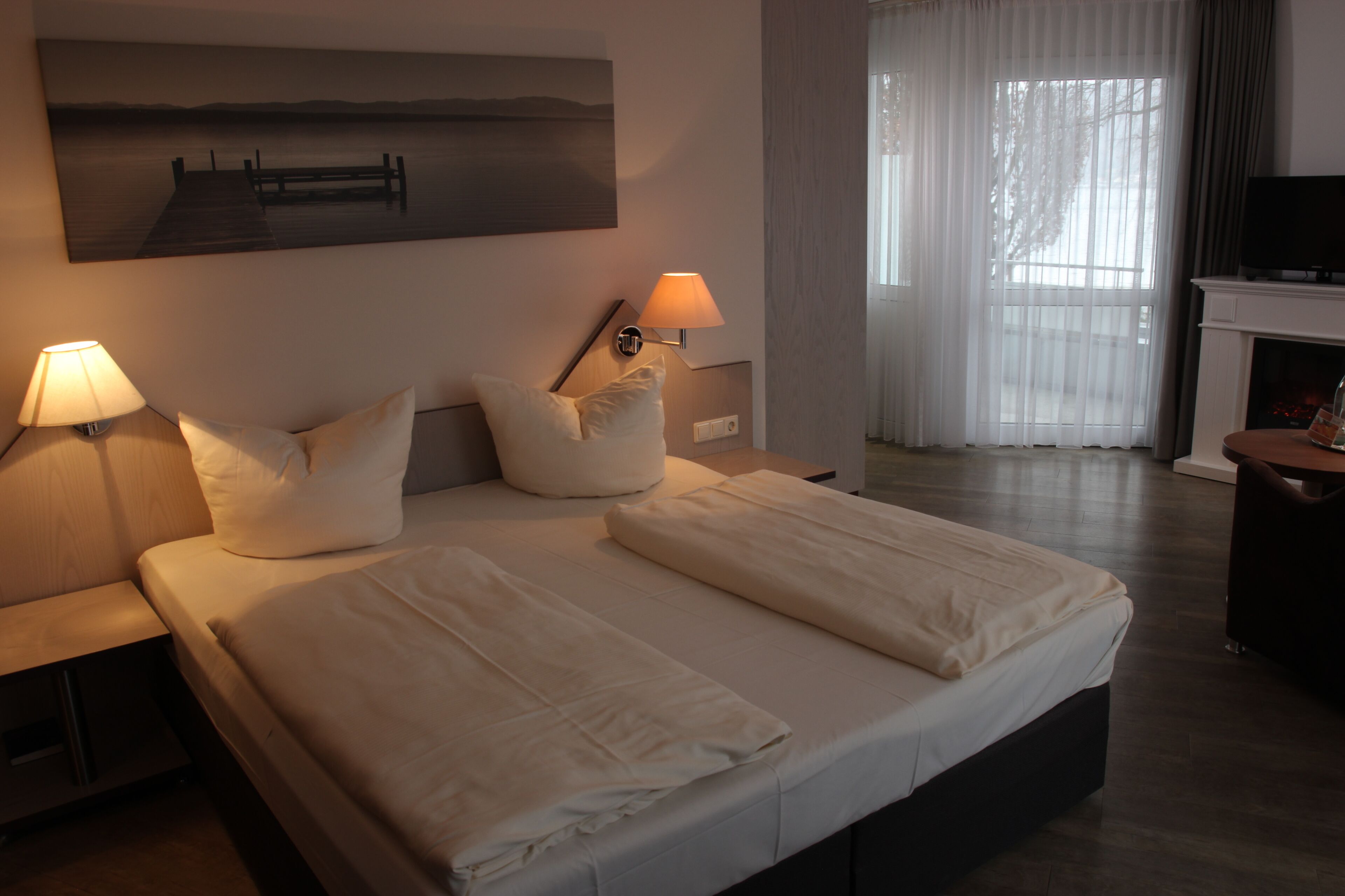 Quarto de hotel com cama, duas luminárias de cabeceira, um quadro, lareira e televisão.