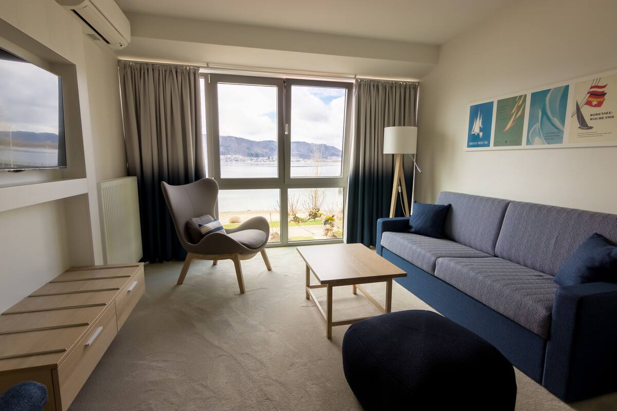 Quarto de hotel moderno com uma janela grande, um sofá, uma poltrona, uma mesinha e uma televisão.