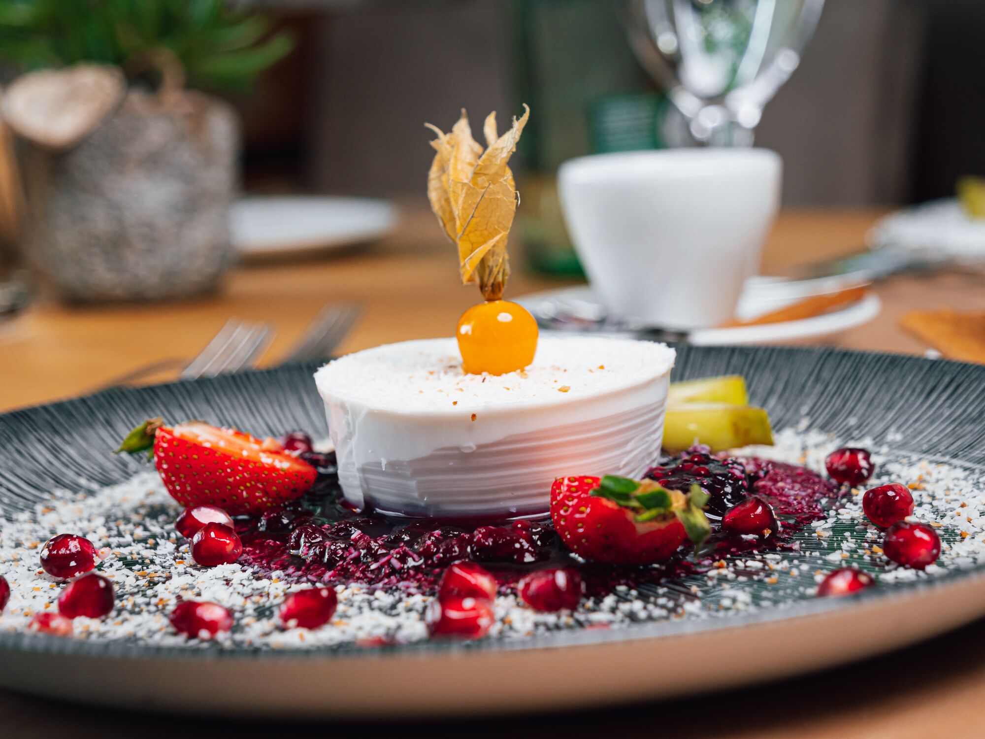 Un dessert à base blanche, surmonté d’une sphère jaune et d’une feuille décorative, le tout entouré de fraises et de baies sur une assiette à texture particulière.