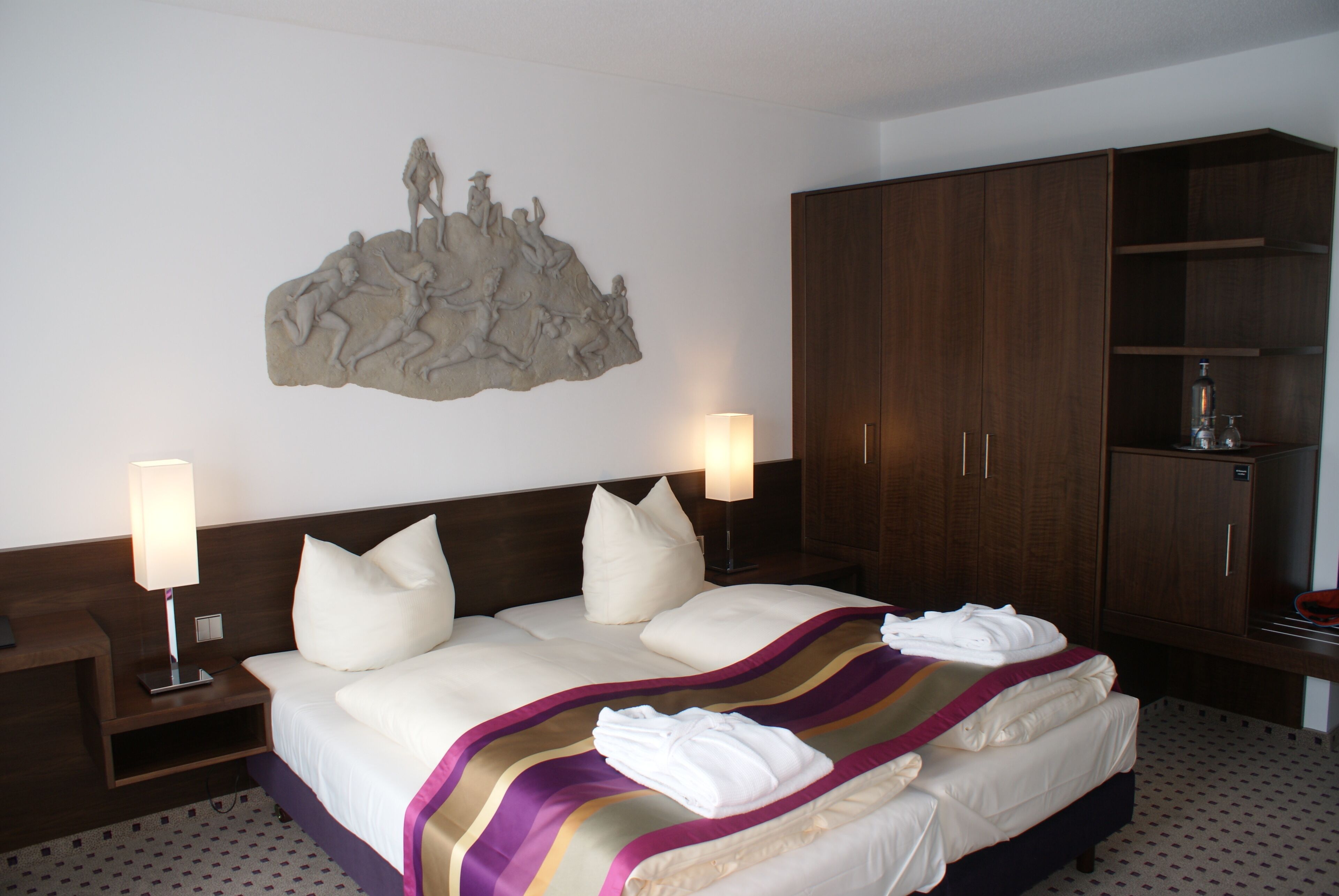 A hotel room with a large bed, two bedside lamps, a wooden wardrobe, and a wall-mounted sculpture.