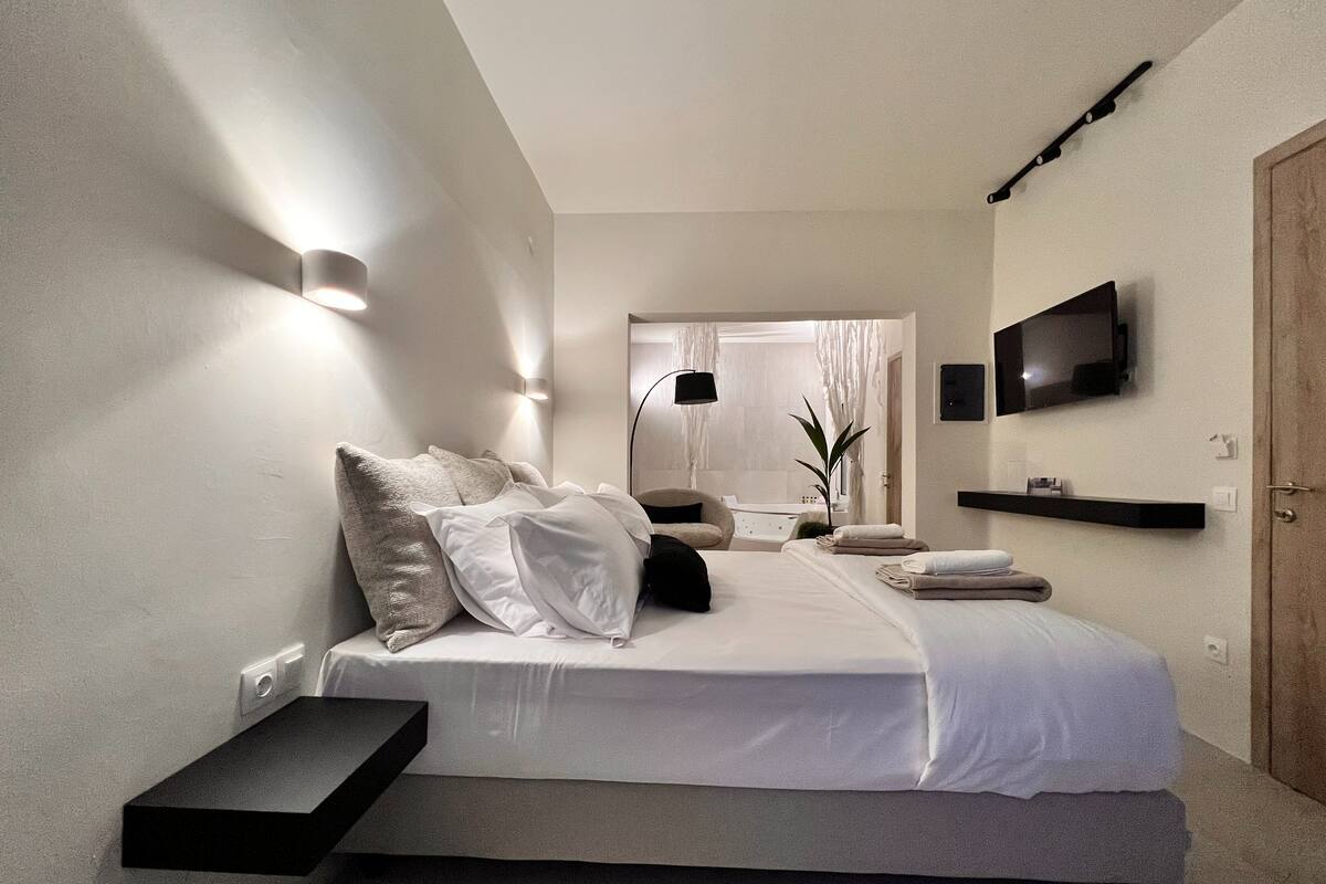 Une chambre moderne avec un lit, des tables de chevet, une télévision et une salle de bain visible à travers une porte ouverte.
