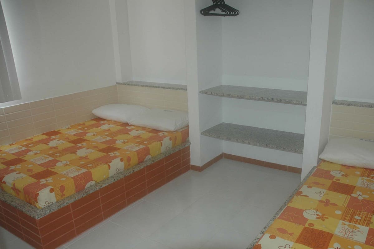 A small, single-room with two beds, each with a quilted blanket. There is a shelf unit with a hanging item above one bed.