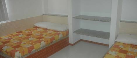 A small, single-room with two beds, each with a quilted blanket. There is a shelf unit with a hanging item above one bed.