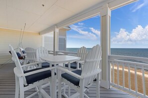 A covered outdoor area with white Adirondack chairs and a table, overlooking a beach.