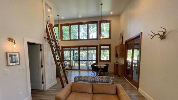 A spacious living area with a large window, a sofa, a dining table, and a ladder leading to an upper level. - Pushin' Time Hideaway at The Retreat at Water's Edge (Tracy City)