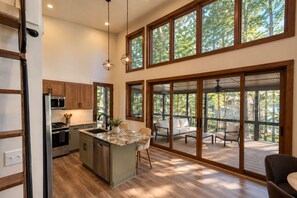 A modern kitchen with a central island, wooden cabinets, and a large sliding glass door leading to an outdoor deck with seating. - Pushin' Time Hideaway at The Retreat at Water's Edge (Tracy City)