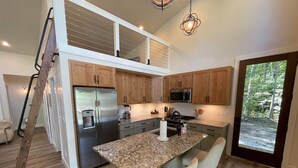 A modern kitchen with a stainless steel refrigerator, a built-in microwave, granite countertops, and wooden cabinets. There is a small island with bar stools and a view of the outdoors through a glass door. - Pushin' Time Hideaway at The Retreat at Water's Edge (Tracy City)