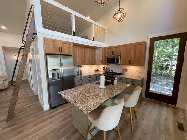 Una cocina moderna con refrigerador de acero inoxidable, microondas integrado, encimeras de granito y armarios de madera. Hay una pequeña isla con taburetes de bar y vistas al exterior a través de una puerta de vidrio.