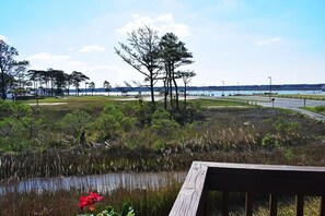 Un paysage côtier avec une terrasse en bois, une étendue d’eau et une côte lointaine parsemée de bâtiments et d’arbres.