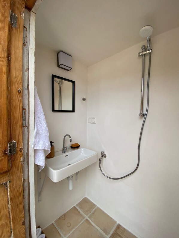 Un baño con lavamanos blanco, un espejo, un cabezal de ducha manual y una puerta de madera.