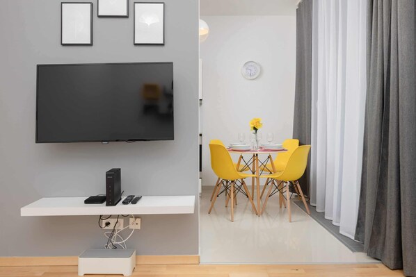 A modern living room with a flat-screen TV, a white console table with electronic devices, a dining area with yellow chairs and a small table, and a gray curtain.