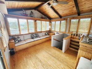A cabin interior with a wooden floor, a built-in bench with cushions, a small kitchen area, and large windows with blinds. - Treehouse- Fire Pit, Basketball Court, Bikes, Kayaks & Casita w/ Game Room (Chincoteague)