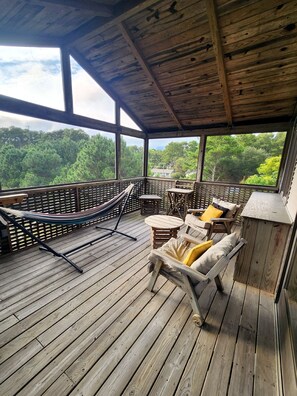 A covered wooden deck with outdoor furniture, including a hammock and chairs, overlooking a forested area. - Treehouse- Fire Pit, Basketball Court, Bikes, Kayaks & Casita w/ Game Room (Chincoteague)