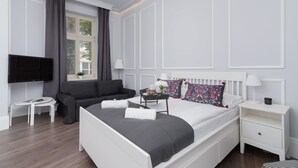 Ein modernes Schlafzimmer mit einem weißen Bett, einem grauen Sofa und einem an der Wand befestigten Fernseher.