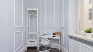 Ein kompakter Arbeitsraum mit weißem Schreibtisch, Stuhl und Regalen, einem Heizkörper und einem Fenster mit einer Pflanze.