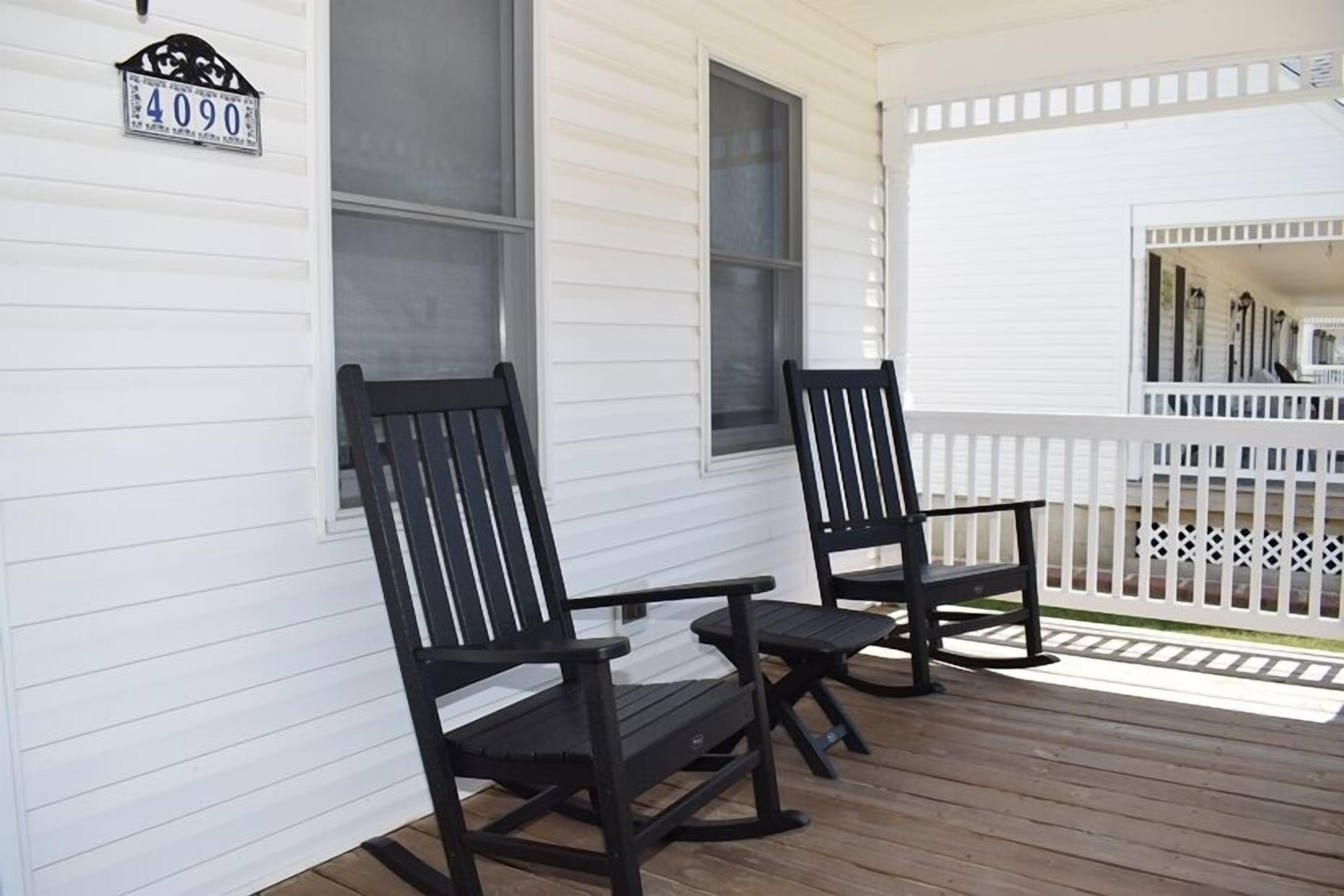 A porch with two rocking chairs, a white fence, and a house number sign.