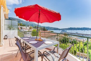 A balcony with a table set for two, a red umbrella, and a view of a marina and hills. - LA MALMAISON II AP4315 By Riviera Holiday Homes (Villefranche-sur-Mer)