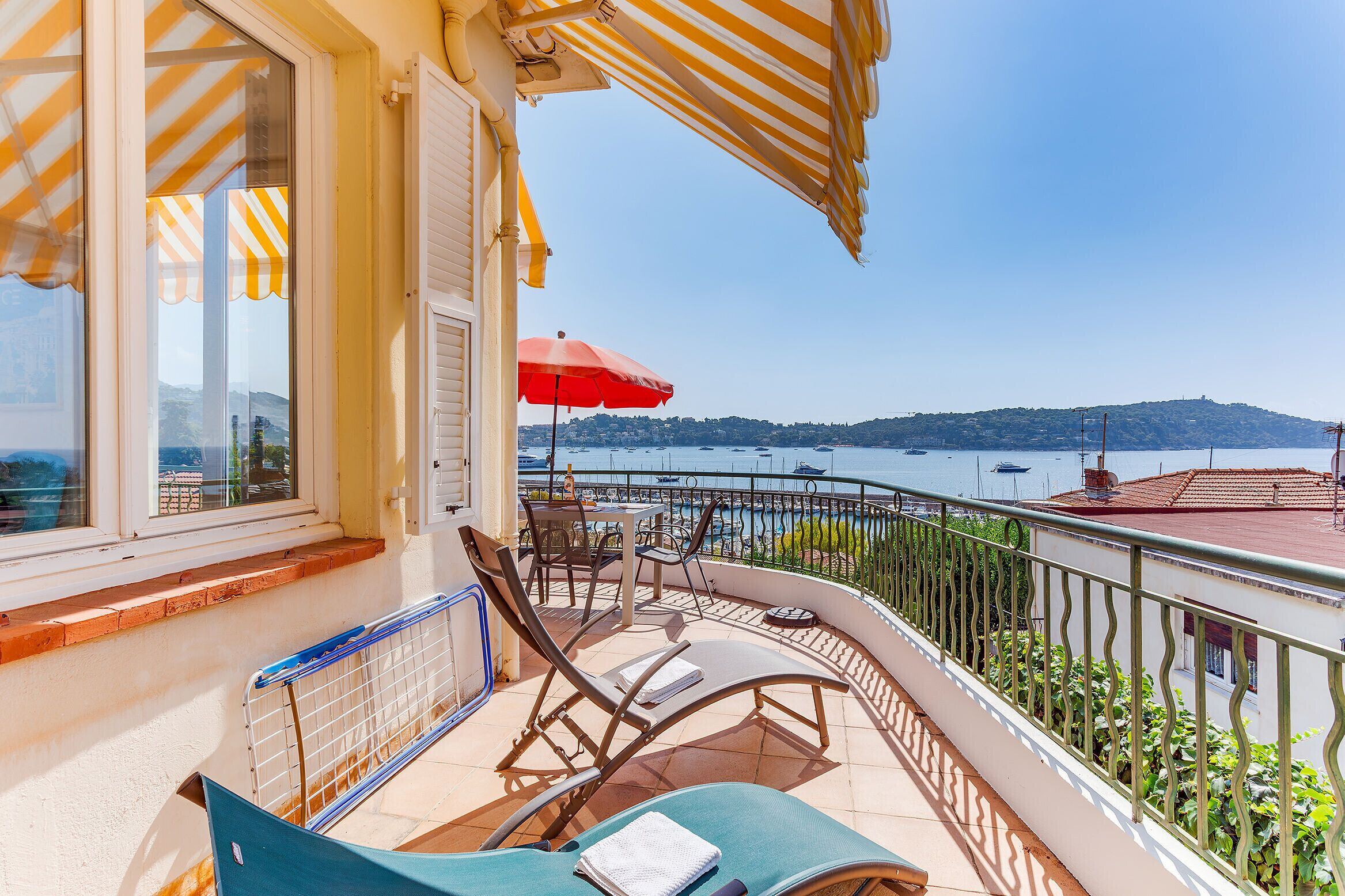 A balcony with a view of the sea, a table with chairs, and a red umbrella.