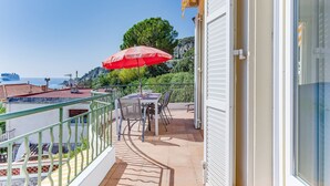 En balkon med et bord og stole, en rød parasol og udsigt til havet og bygninger.