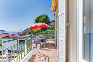 A balcony with a table and chairs, a red umbrella, and a view of the sea and buildings. - LA MALMAISON II AP4315 By Riviera Holiday Homes (Villefranche-sur-Mer)
