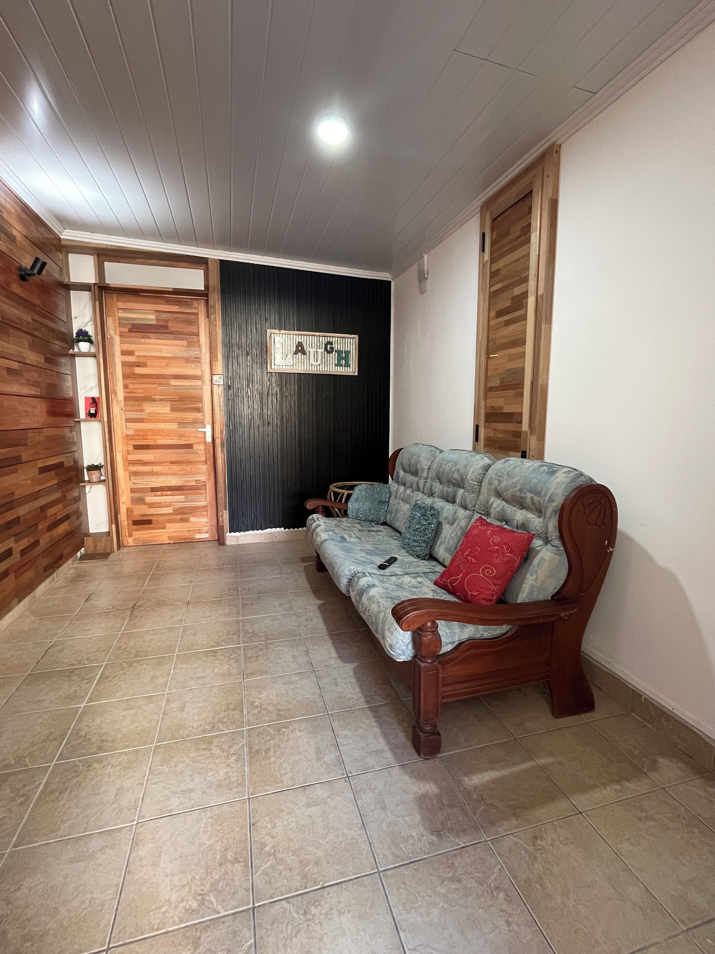 Una habitación con puerta de madera, pared negra con un cartel que dice 'LAUGH', un banco de madera y piso de baldosas.