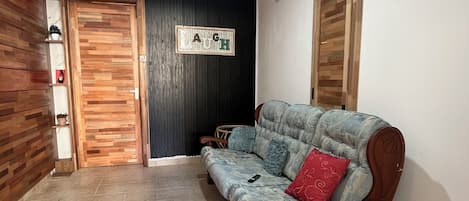 Une pièce avec une porte en bois, un mur noir orné d’un panneau indiquant le mot « RIRE », un banc en bois et un sol carrelé.