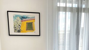 Un área de comedor con una mesa azul aguamarina y dos sillas, una obra de arte enmarcada en la pared y una ventana con cortinas traslúcidas.