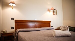 A hotel room with a wooden headboard, a bed with a quilted bedspread, two wall-mounted lamps, a bedside table with a telephone, and a rolled towel on the bed. - Bagno Santo Residence (Manciano)