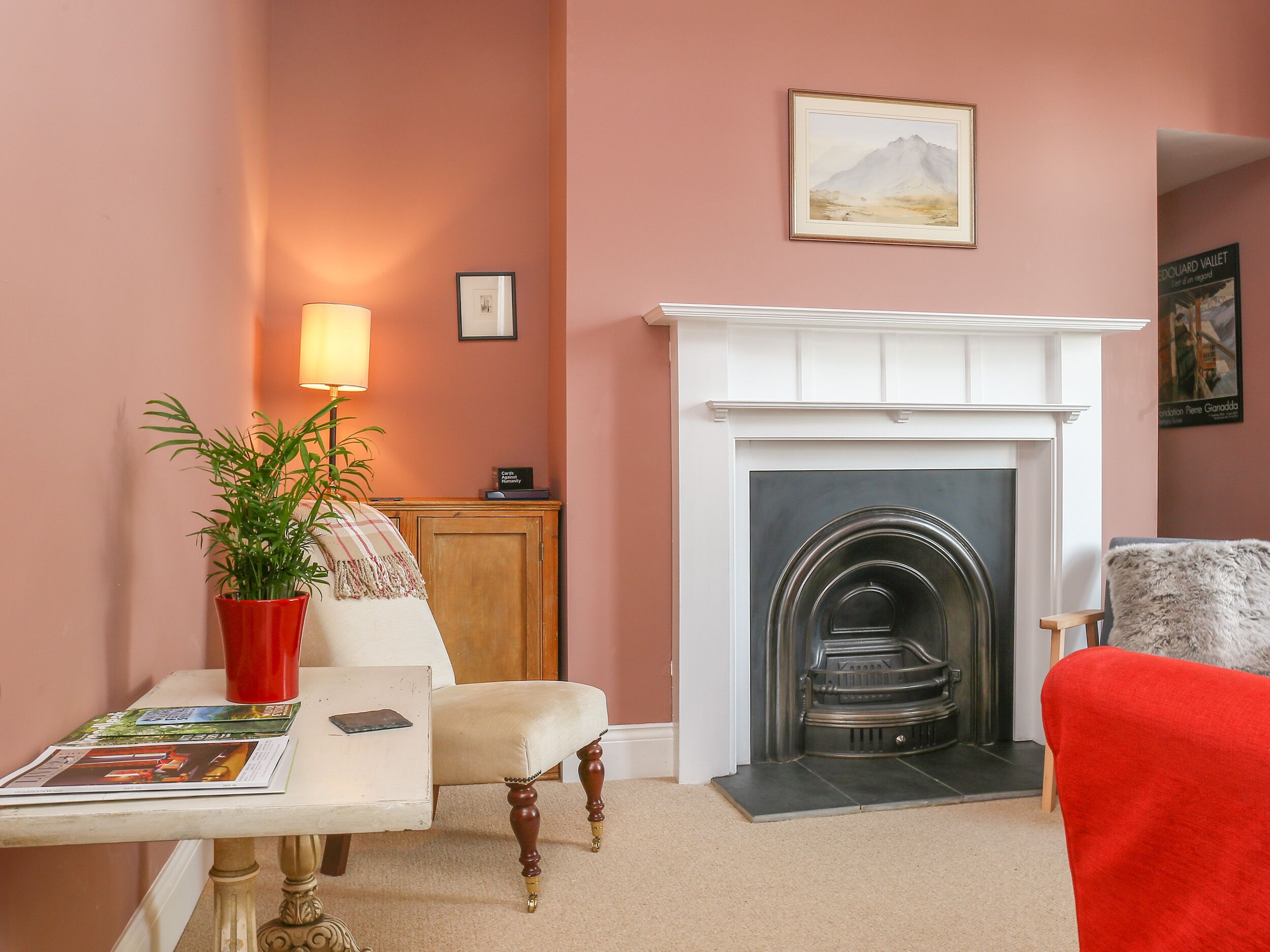 A cozy living room with a fireplace, a red plant, a white armchair, and a red chair.