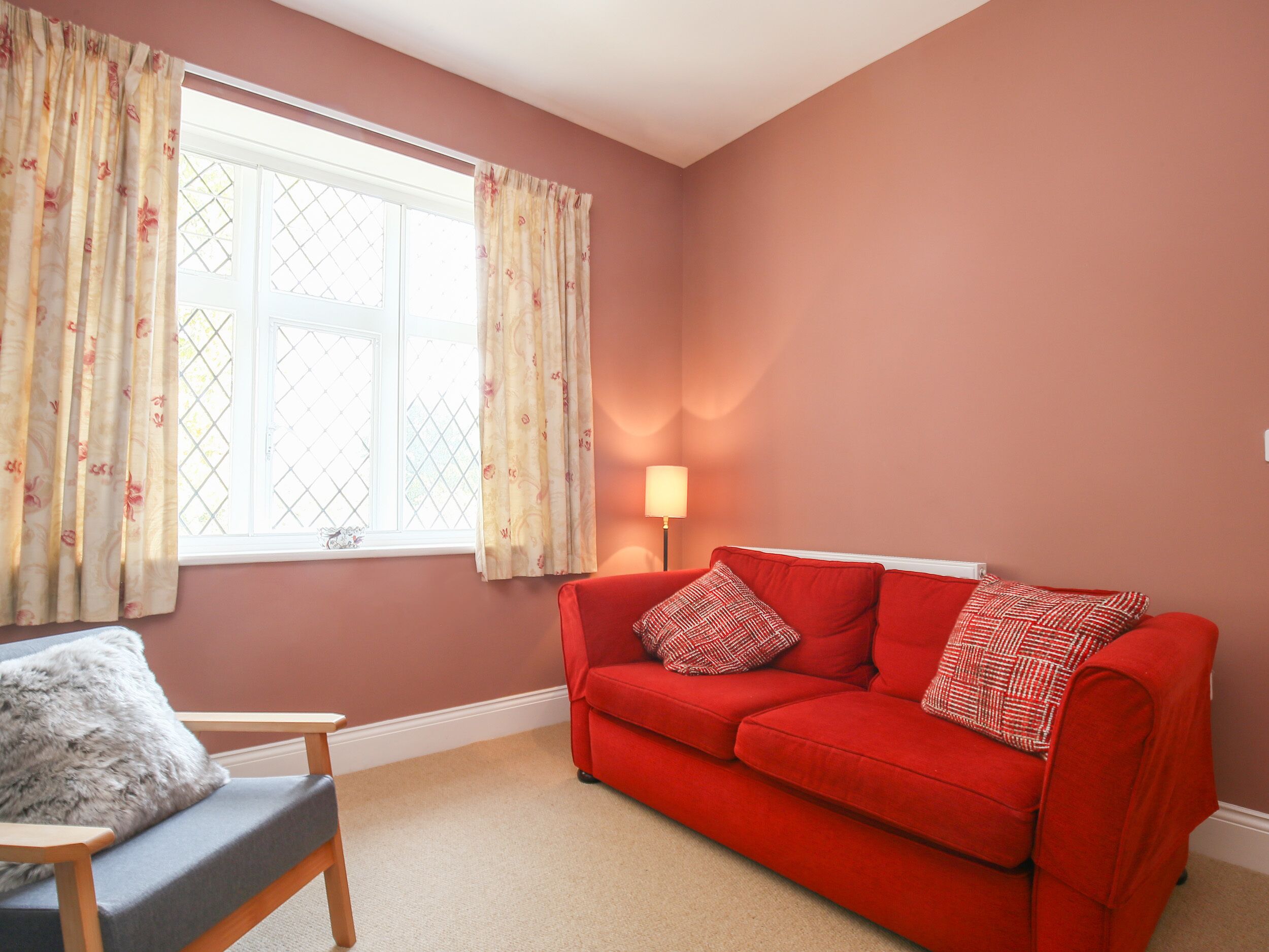 A living room with a red sofa, a grey armchair, and a window with floral curtains.
