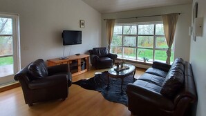A spacious living room with a wooden ceiling, a large window with curtains, leather sofas, and a flat-screen TV. - Hótel Skálholt (Bláskógabyggd)