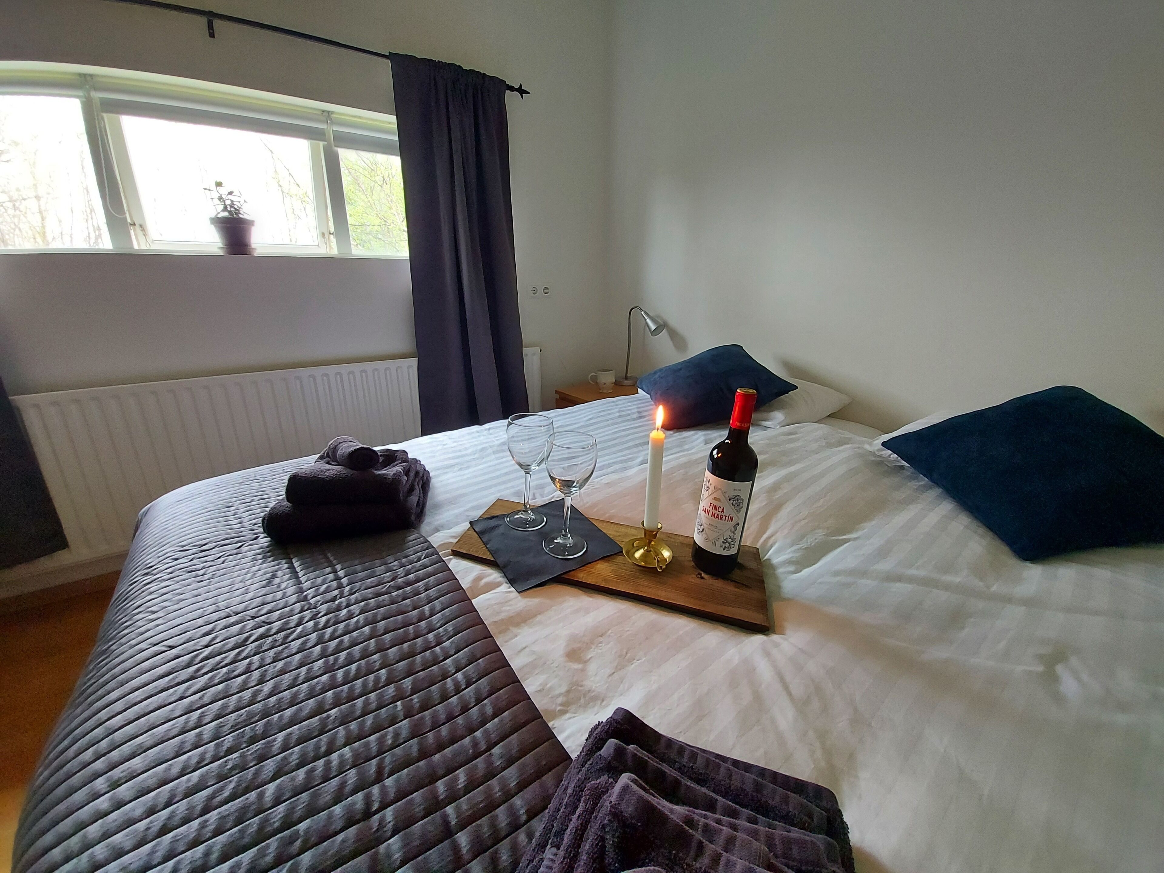 Una cama bien hecha con una botella de vino, copas y una vela sobre una mesita de noche.