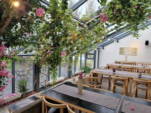 An indoor dining area with tables and chairs, a greenhouse with climbing plants and flowers, and a view through large windows. - Hótel Skálholt (Bláskógabyggd)