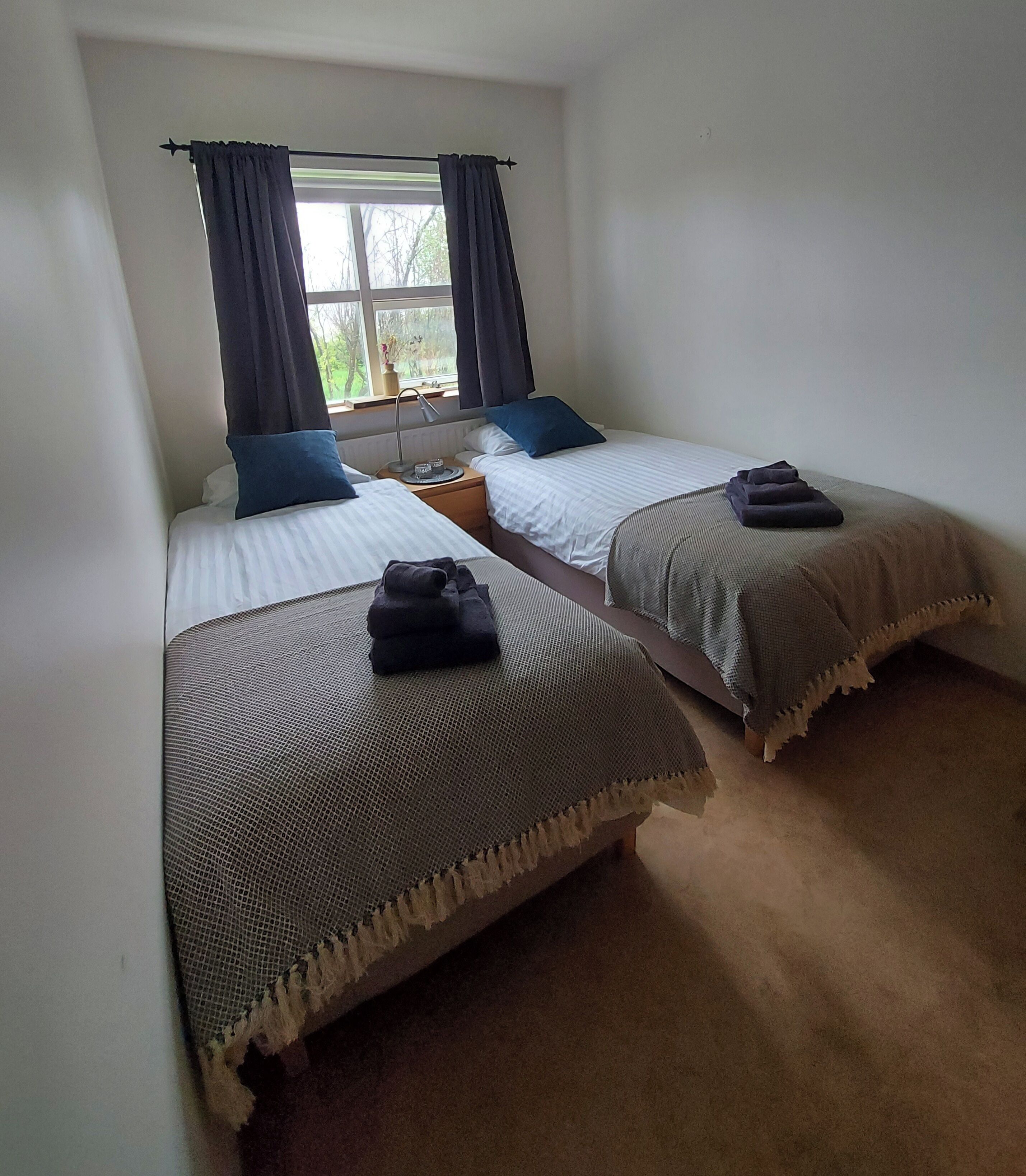 A room with two beds, each with a pillow and a folded towel. There is a window with curtains, a nightstand with a lamp, and a vase with flowers.