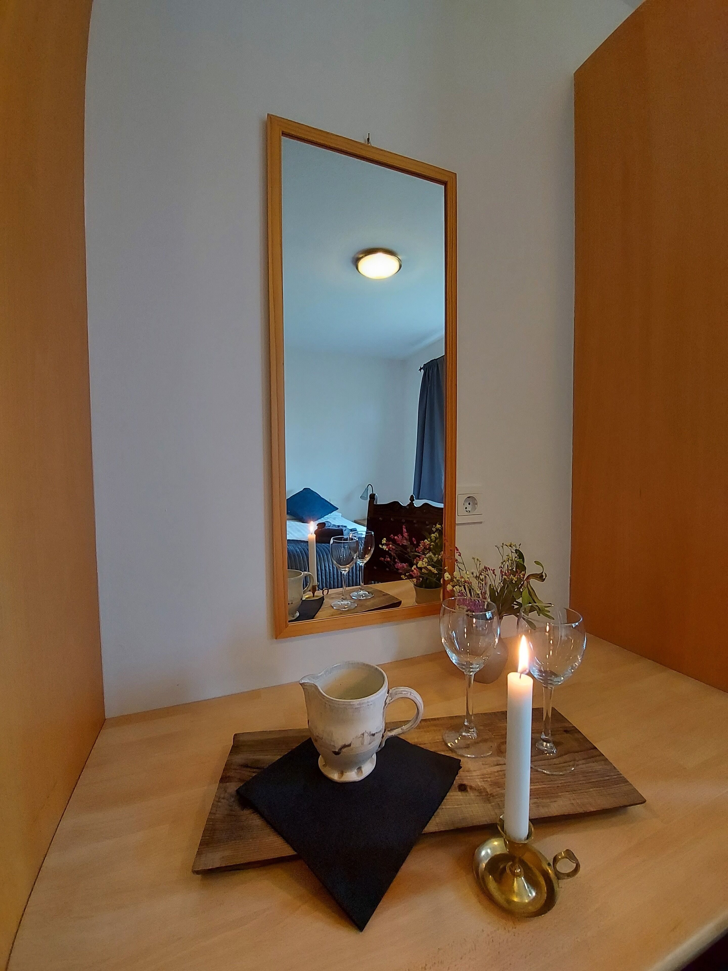 Una mesa de madera con una jarrón blanco, una vela encendida y dos copas de vino sobre un mantel negro. Un espejo refleja una habitación con una cama y una ventana.