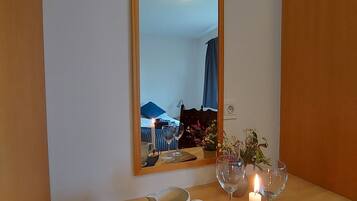 Una mesa de madera con una jarrón blanco, una vela encendida y dos copas de vino sobre un mantel negro. Un espejo refleja una habitación con una cama y una ventana.