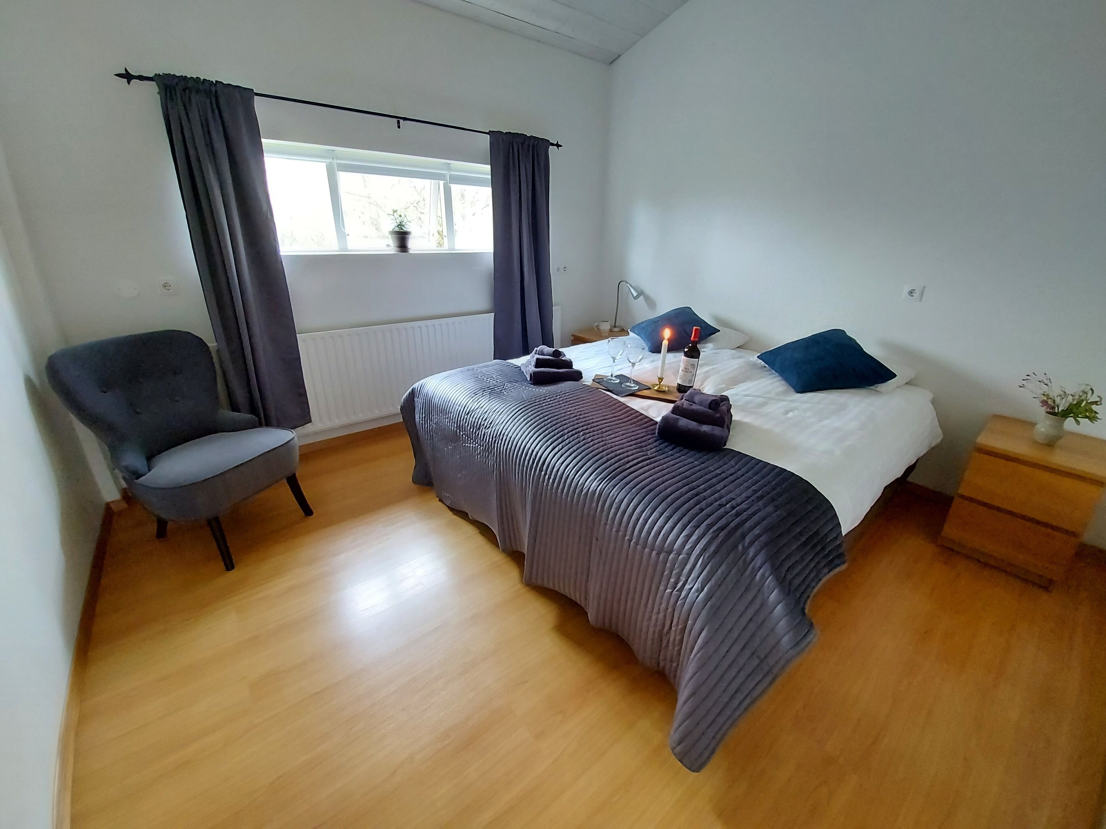 Ein Schlafzimmer mit Bett, Sessel, Nachttisch mit Lampe und einem Fenster mit Vorhängen.
