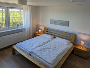 Ein Schlafzimmer mit einem hölzernen Bett, weißer Bettwäsche, einem fenster mit Vorhängen, einer Heizung und einem an der Wand befestigten Kunstwerk.