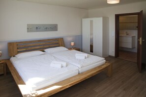 A bedroom with a wooden bed, white bedding, a nightstand, a bedside lamp, a wardrobe, and a bathroom visible through an open door.