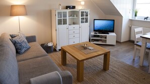 Ein modernes Wohnzimmer mit einem grauen Sofa, einem Holztisch, einem Fernseher und einer weißen Vitrine mit Glastüren.
