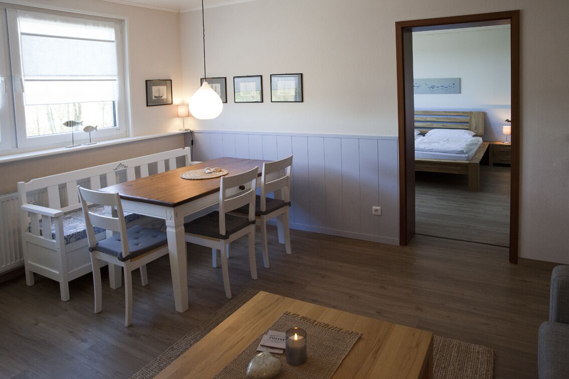 Ein gemütliches Zimmer mit einem Holzesstisch, weißen Stühlen, einem kleinen Teppich und einem Nachttisch mit Lampe.