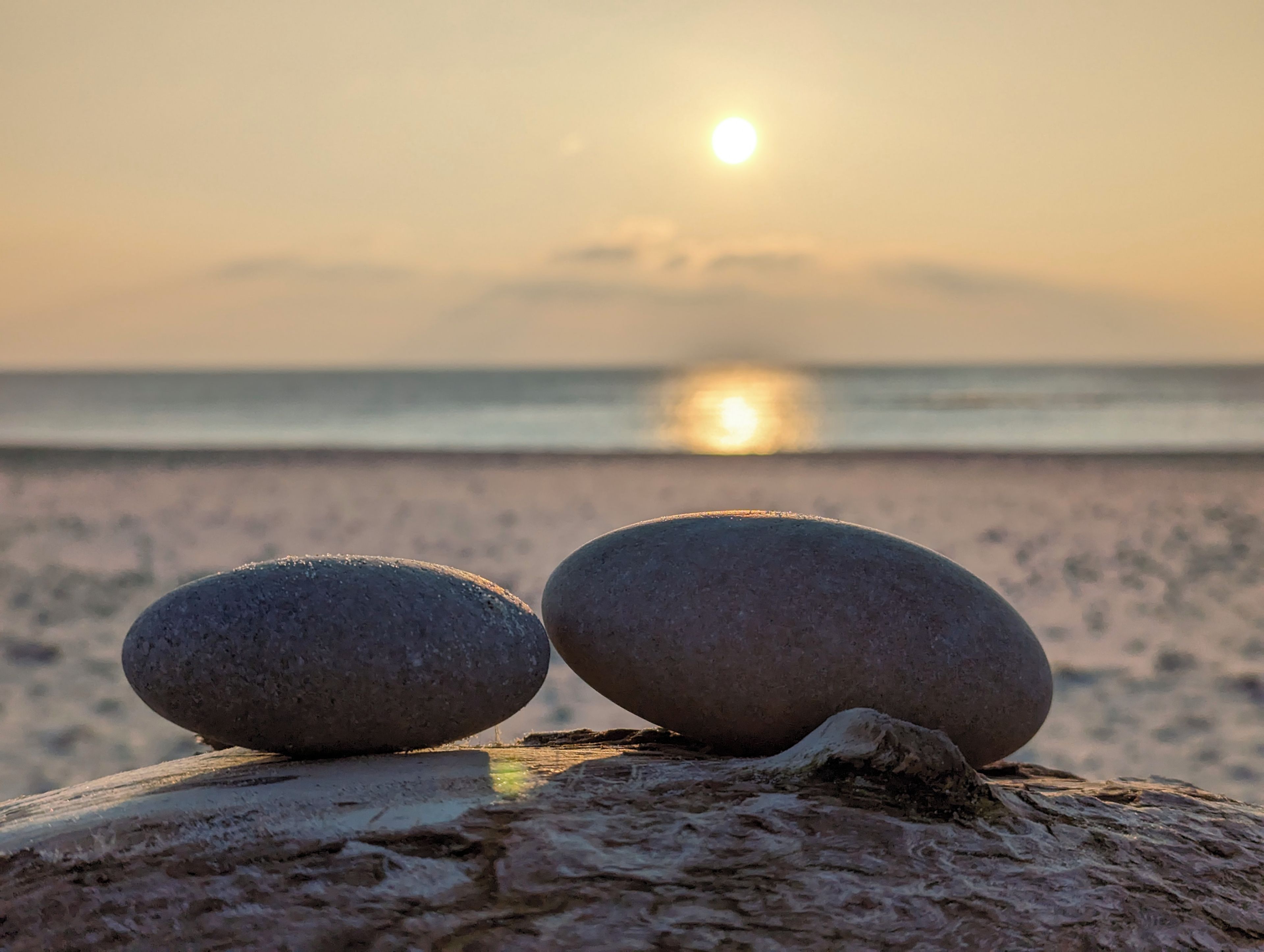 Twee gladde stenen balanceren op een houten ondergrond, met een rustige zee en een ondergaande zon op de achtergrond.