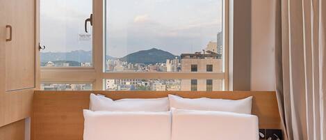 A hotel room with a bed, a window with a view of a cityscape, and a wooden wardrobe.