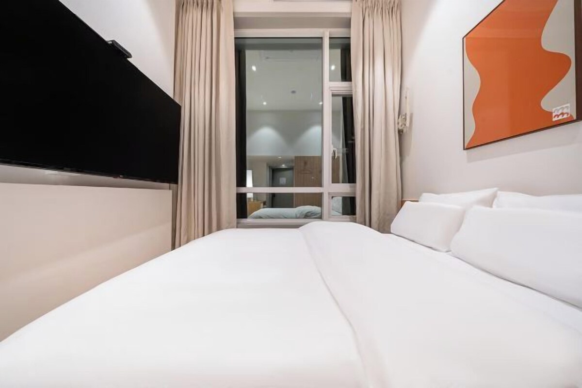 Ein Hotelzimmer mit einem Bett, einem an der Wand befestigten Fernseher, einem Fenster mit Vorhängen und einem Wandbild.
