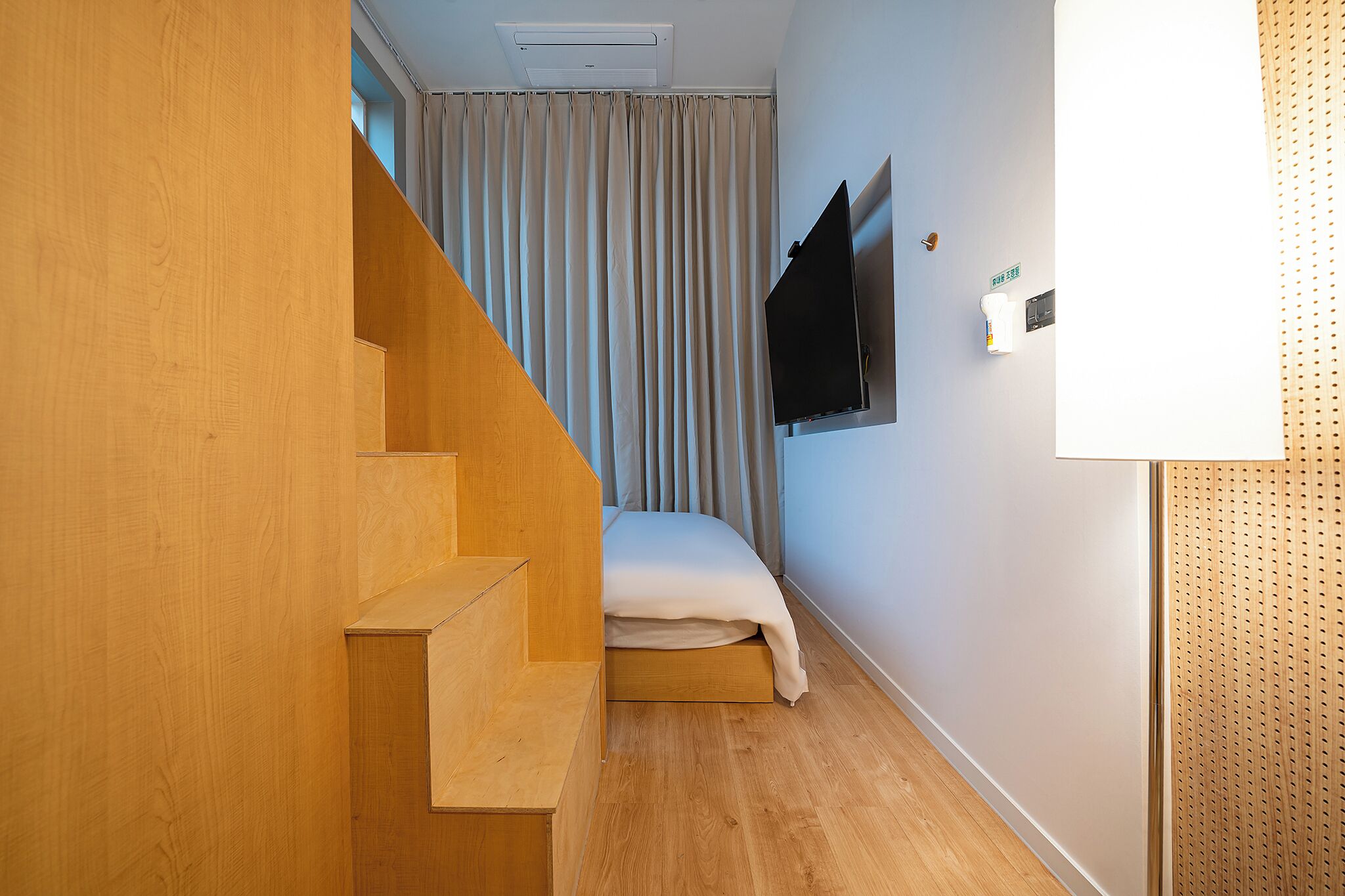 A room with a wooden staircase, a bed with a white mattress, a mounted TV, and a window with curtains.