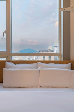Un lit avec une literie blanche, une fenêtre donnant sur un paysage urbain et une montagne, et une porte avec une poignée.